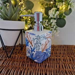 May include: A square ceramic soap dispenser with a chrome pump. The dispenser features a blue and brown coral reef pattern. It is placed on a woven surface.
