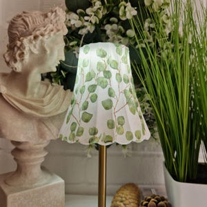 May include: A small lamp with a white lampshade decorated with a green eucalyptus leaf design. The lamp has a gold-coloured base. The lamp is placed in front of a white bust and greenery.