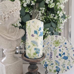 Pode incluir: Uma vela de pilar branca decorada com desenhos florais azuis e roxos, colocada em um castiçal de madeira escura. Um prato floral correspondente e um busto clássico estão no fundo, com uma coroa de vegetação.