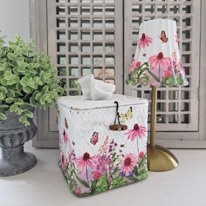 Può includere: Un portatovaglioli bianco con un motivo floreale con fiori rosa, farfalle e api. La scatola ha una chiusura a levetta in legno e un fazzoletto che sporge. È visibile anche un paralume abbinato con lo stesso motivo.