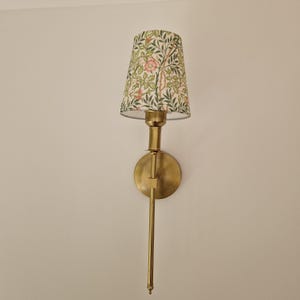Könnte beinhalten: Eine Wandleuchte mit Messing-Finish. Der Lampenschirm hat ein Blumenmuster mit grünen Blättern und roten Blüten auf cremefarbenem Hintergrund. Die Lampe hat einen langen, schlanken Arm und einen kreisförmigen Sockel.