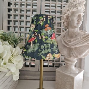 May include: A small table lamp with a dark blue lampshade featuring a botanical print of mushrooms, berries, and ferns. The lamp has a gold-colored stem and a scalloped edge on the shade. A white bust is in the background.