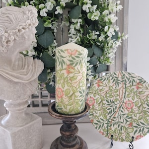 May include: A cream-colored pillar candle with a floral design featuring pink roses and green leaves. The candle sits on a dark wooden candlestick. A matching decorative plate with the same floral pattern is also visible.