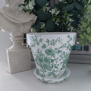 May include: A white ceramic flower pot with a green botanical print featuring intricate floral and vine patterns. The pot includes a matching circular saucer base and has a wide, flared rim. The design covers the exterior surface of the pot.