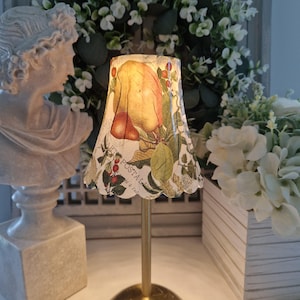 May include: A small decorative lamp with a gold-coloured base and a lampshade featuring a fruit and botanical design. The lampshade has a scalloped edge and text that reads "ART POSTAL". The lamp is placed near a white bust and floral arrangements.