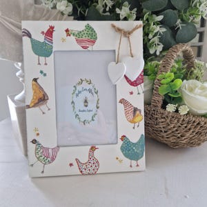 Può includere: Cornice fotografica rettangolare bianca decorata con illustrazioni colorate di galline e uccelli. La cornice ha un centro vuoto per una foto e due decorazioni bianche a forma di cuore. Il testo "Apley Ever After" è visibile.