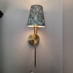 Pode incluir: Um candeeiro de parede com braço em latão escovado e base circular. O abajur cónico apresenta um padrão botânico detalhado com flores azuis e folhagem verde sobre um fundo escuro, emitindo uma luz quente.