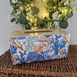 Puede incluir: Caja de pañuelos rectangular con tapa de madera clara y fondo blanco. Los lados están decorados con diseños de arrecifes de coral azules, marrones y color coral. La caja de pañuelos está sobre una superficie de mimbre tejido.