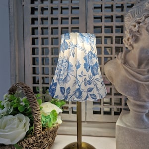 Puede incluir: Una pequeña lámpara de mesa con una pantalla plisada con un estampado floral azul sobre un fondo blanco. La lámpara tiene una base y un tallo de color latón. Una cesta de rosas blancas y vegetación está en primer plano.