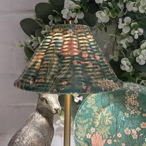 May include: A table lamp with a gold-toned metal base and a pleated, dark green fabric shade featuring a floral pattern with pink tulips. Beside the lamp sits a metallic, textured rabbit figurine and a round, green floral-patterned decorative plate.