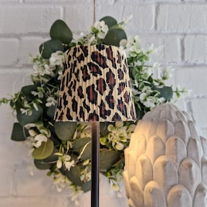 Può includere: Un piccolo paralume con un motivo leopardato marrone e nero. Il paralume è plissettato e poggia su un supporto in metallo scuro. Sullo sfondo una ghirlanda decorativa con fiori bianchi e foglie verdi.