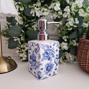 Puede incluir: Dispensador de jabón cuadrado con base blanca y estampado floral azul. Tiene una bomba plateada y se encuentra sobre un fondo de follaje verde y flores blancas. Aproximadamente 10 cm de altura.