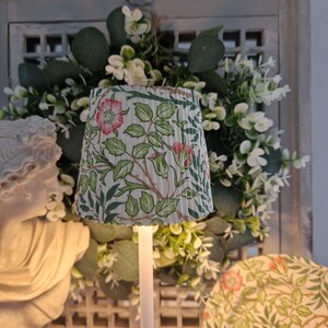 May include: A small, pleated lampshade with a floral design of pink roses and green leaves on a white base. The lampshade is set against a backdrop of greenery and a decorative wreath, creating a vintage aesthetic.