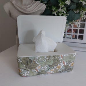 May include: A rectangular tissue box holder with a hinged white lid and a base featuring a repeating botanical pattern of sage green leaves and small yellow flowers on a cream background, with a single white tissue protruding from the top opening.