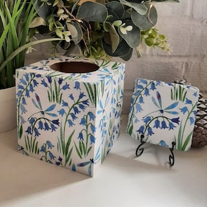 Könnte beinhalten: Ein quadratischer Taschentuchspender und ein Untersetzer mit einem Blaubeeren- und Libellen-Design. Der weiße Hintergrund ist mit Blaubeerenblüten und grünen Stielen verziert. Der Untersetzer wird auf einem schwarzen Metallständer präsentiert.