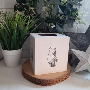 May include: A square, off-white tissue box with a circular opening on top. A black and white illustration of a bear is on the front. The box sits on a round, wooden base. A potted plant and decorative star are in the background.