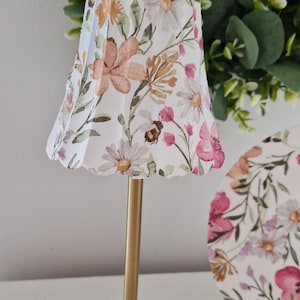 May include: A gold table lamp with a white lampshade featuring a floral pattern with pink, orange, and green flowers. The lampshade has a scalloped edge.