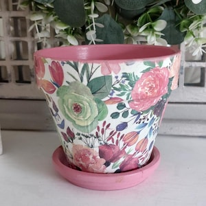 May include: A tapered flower pot with a matching saucer, both featuring a solid pink rim and base. The exterior of the pot is decorated with a colourful floral pattern of pink, green, and orange flowers and leaves against a white background.