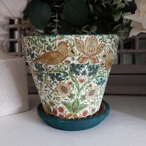 May include: A tapered terracotta plant pot featuring a detailed botanical pattern with orange birds, green foliage, yellow flowers, and small red strawberries. The rim and the matching circular saucer are painted in a solid dark teal green colour.