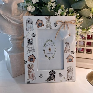 Può includere: Cornice bianca decorata con illustrazioni ad acquerello di varie razze di cani, casette per cani, impronte di zampe e ossa. La cornice ha un fiocco di spago e due ornamenti a forma di cuore. Il testo "Ashley Ever After" è visibile.