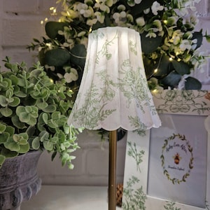 Op de afbeelding: Een geplooide lampenkap met een geschulpte onderrand, voorzien van een witte achtergrond en een groene botanische print van bloemen en bladeren. De kap is gemonteerd op een dunne, cilindrische messingkleurige metalen standaard.