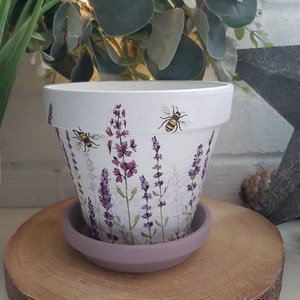 May include: White ceramic plant pot with a lavender and bee design. The pot features purple lavender flowers and two bees. It sits on a matching lavender-coloured saucer, ideal for indoor or outdoor plants.