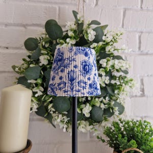 Puede incluir: Una pantalla de lámpara floral azul y blanca con un diseño plisado. La pantalla de la lámpara está colocada contra una pared de ladrillos blancos, con una corona floral verde y blanca en el fondo. También se ven una vela color crema y una pequeña planta en maceta.