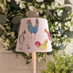 Può includere: Una lampada bianca con un paralume conico decorato con illustrazioni colorate di galline e galli. La lampada è ambientata su uno sfondo di verde e un muro di mattoni bianchi, creando un'estetica da fattoria.