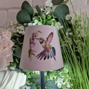 May include: A small, off-white lampshade featuring a colorful hummingbird design. The hummingbird is depicted with outstretched wings, feeding from purple flowers. The lampshade is surrounded by greenery and a white brick background.