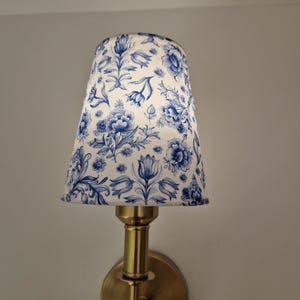 May include: A wall-mounted lamp with a brass base and a lampshade featuring a blue and white floral pattern. The lampshade is cone-shaped, and the floral design includes various flower and leaf motifs.