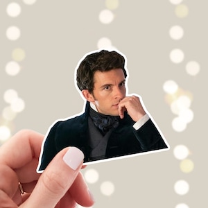 May include: A sticker featuring a portrait of a man with dark hair, wearing a dark blue suit and a white shirt with a black bow tie. He is looking to the side and has his hand on his chin.