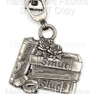 May include: Silver-toned zipper pull featuring a stack of books with the words "Smut" and "Slut" on the spines. The design includes a rose and a book with a chili pepper. A metal ring connects the pull to the zipper.