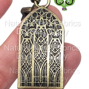 May include: A gold-toned metal charm featuring a detailed design of a stained glass window with a geometric pattern. The charm has a loop at the top for attaching to a necklace or bracelet.