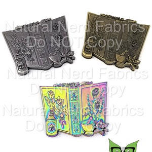 May include: A rainbow iridescent enamel pin depicting an open book with a mortar and pestle, a bottle of potion, and floral illustrations. The pin is a whimsical and colourful representation of herbalism and magic.