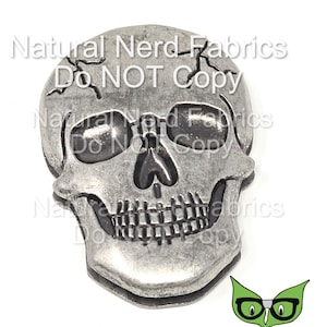 May include: A silver skull charm with a cracked surface and black eye sockets. The skull is a decorative embellishment for crafts and jewelry making.