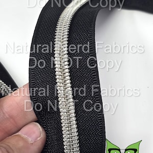 May include: Close-up of a black zipper with a silver metal chain. The zipper is folded in half, showing the inside and outside of the zipper tape. The zipper is made of a durable material and has a smooth finish.