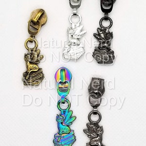 Puede incluir: Cinco tiradores de cremallera con un amuleto de gato leyendo un libro. Los amuletos están en diferentes acabados: plata, negro, oro, iridiscente y gris oscuro.