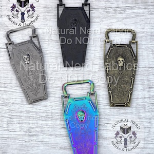May include: Four coffin-shaped metal hardware pieces in black, silver, bronze, and iridescent finishes. Each features a skull and floral design, with a handle at the top. The text "Natural Nerd Fabrics & Hardware" is visible.