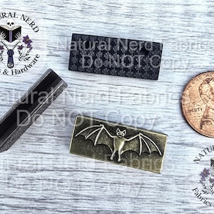 May include: An assortment of metal hardware pieces, including a dark gray rectangular piece with a grid pattern, a bronze-colored rectangle with a bat design, and a U-shaped piece. A US penny is included for size reference. The text "Natural Nerd Fabrics & Hardware" is visible.