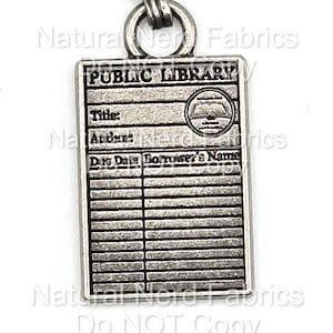 May include: A rectangular, silver-toned pendant featuring a detailed design of a library card. The pendant includes the words "PUBLIC LIBRARY", along with spaces for title, author, due date, and borrower's name. A small, circular library logo is also present.