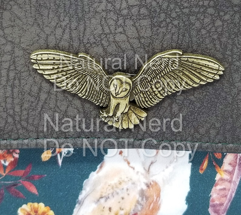 A bronze-toned metal owl magnetic snap with detailed wings and a realistic owl design. The snap is on a brown textured surface.
