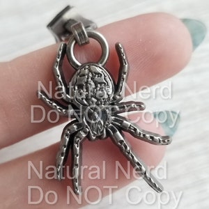 May include: A silver spider charm with a detailed design. The spider has eight legs and a round body with intricate details. The charm is attached to a silver chain.