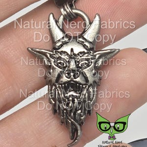 May include: Silver-toned pendant featuring a detailed depiction of a demon's face with horns, fangs, and a beard. The pendant is attached to a silver chain.