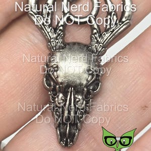 May include: Silver-toned metal charm featuring a detailed skull with antlers. The charm has a ring at the top for attaching to a necklace or bracelet. The charm is a decorative element for jewelry making.