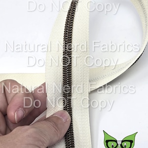 May include: A close-up of a white zipper tape with a brown metal zipper slider. The zipper tape is folded in half and the zipper slider is visible in the center of the image. The text "Natural Nerd Fabrics Do Not Copy" is visible in the background.