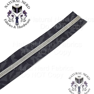 May include: A close-up of a black zipper with a silver metal coil. The zipper tape has a dark pattern. The image includes the Natural Nerd Fabrics & Hardware logo with a skull, book, and zipper.