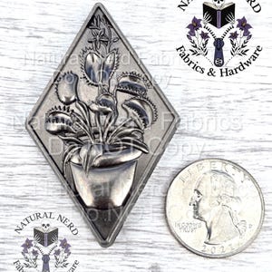 Puede incluir: Un dije de metal plateado en forma de diamante, con un relieve detallado de una planta carnívora Venus atrapamoscas en una maceta. El dije está junto a una moneda para comparar el tamaño. El logotipo de Natural Nerd Fabrics & Hardware es visible.