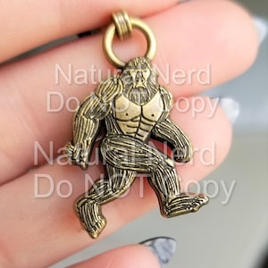 May include: A bronze-colored metal charm of a Bigfoot walking, with a detailed design and a loop at the top for attaching to a necklace or keychain.