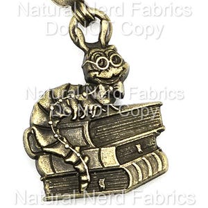 May include: A bronze-colored charm featuring a cartoonish caterpillar wearing glasses and a bow tie, perched atop a stack of books. The charm is attached to a chain and has a detailed, vintage aesthetic. The words "Natural Nerd Fabrics Do NOT Copy" are visible.