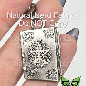 May include: Silver-toned book charm with a five-pointed star design and intricate details. The charm is attached to a silver chain. The text "Natural Nerd Fabrics Do NOT Copy" is visible in the background.